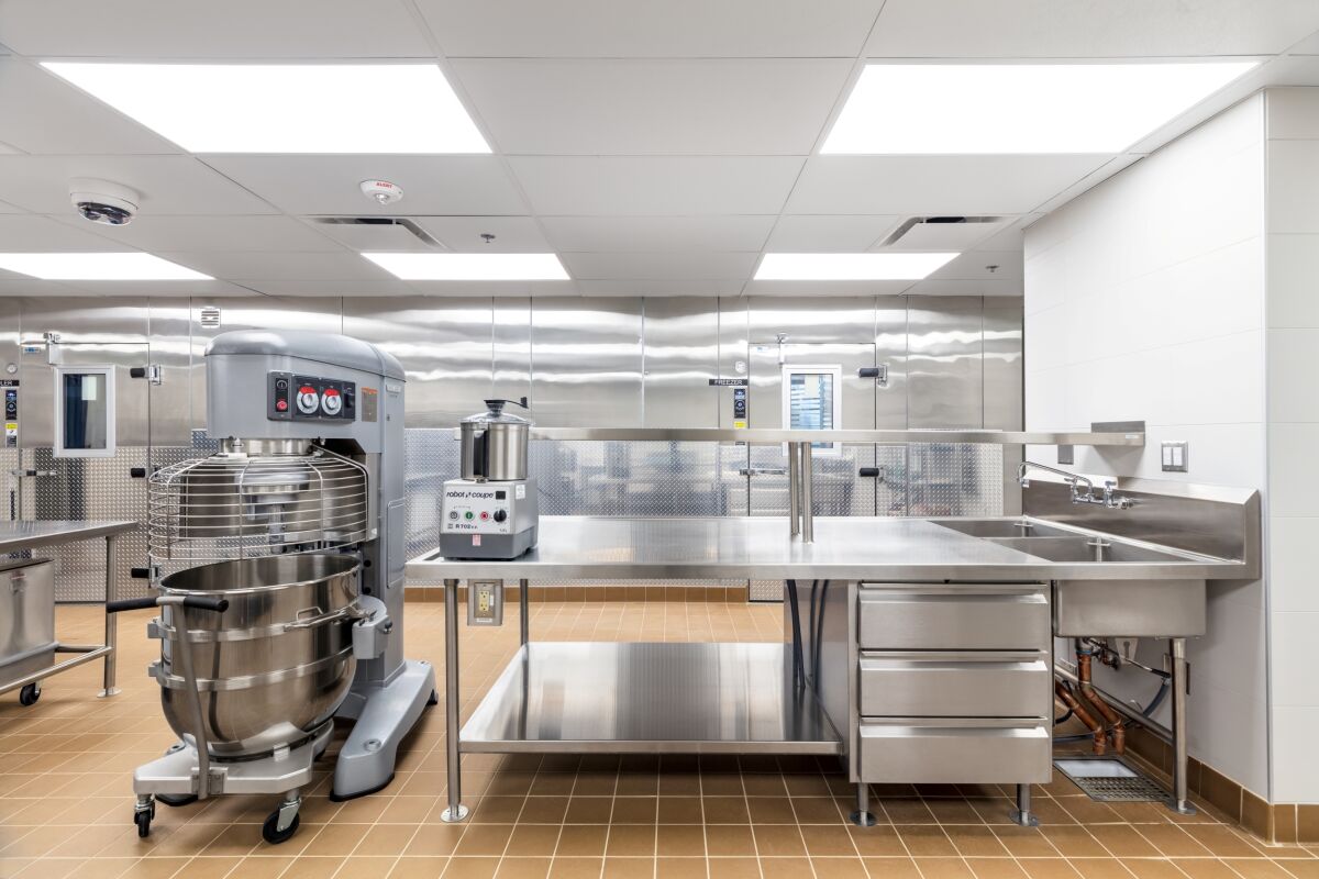 Commercial production kitchen with a large industrial mixer, stainless‑steel prep table with storage drawers, triple‑compartment sink, tile flooring, and diamond‑plate walk‑in cooler doors along the back wall.
