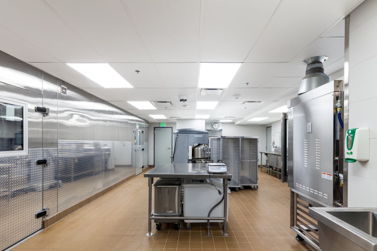 Commercial production kitchen with stainless‑steel walk‑in cooler doors, central prep table, large industrial mixers, rolling sheet‑pan racks, tile flooring with floor drains, and a suspended ceiling with recessed lighting.