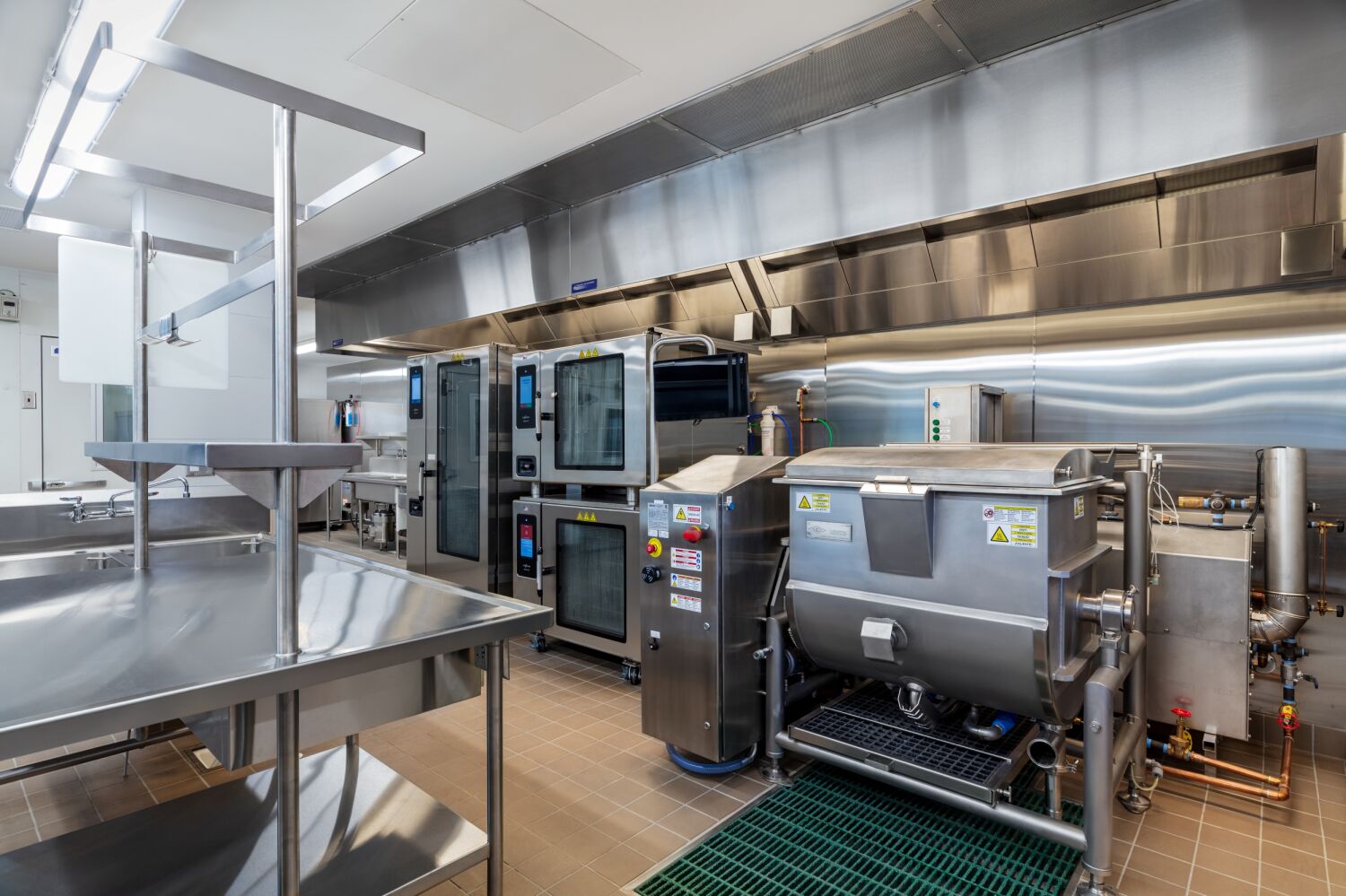 Commercial production kitchen with stainless‑steel equipment including convection ovens, a tilt skillet, prep tables, ventilation hood, tile flooring, and wall‑mounted utility connections.