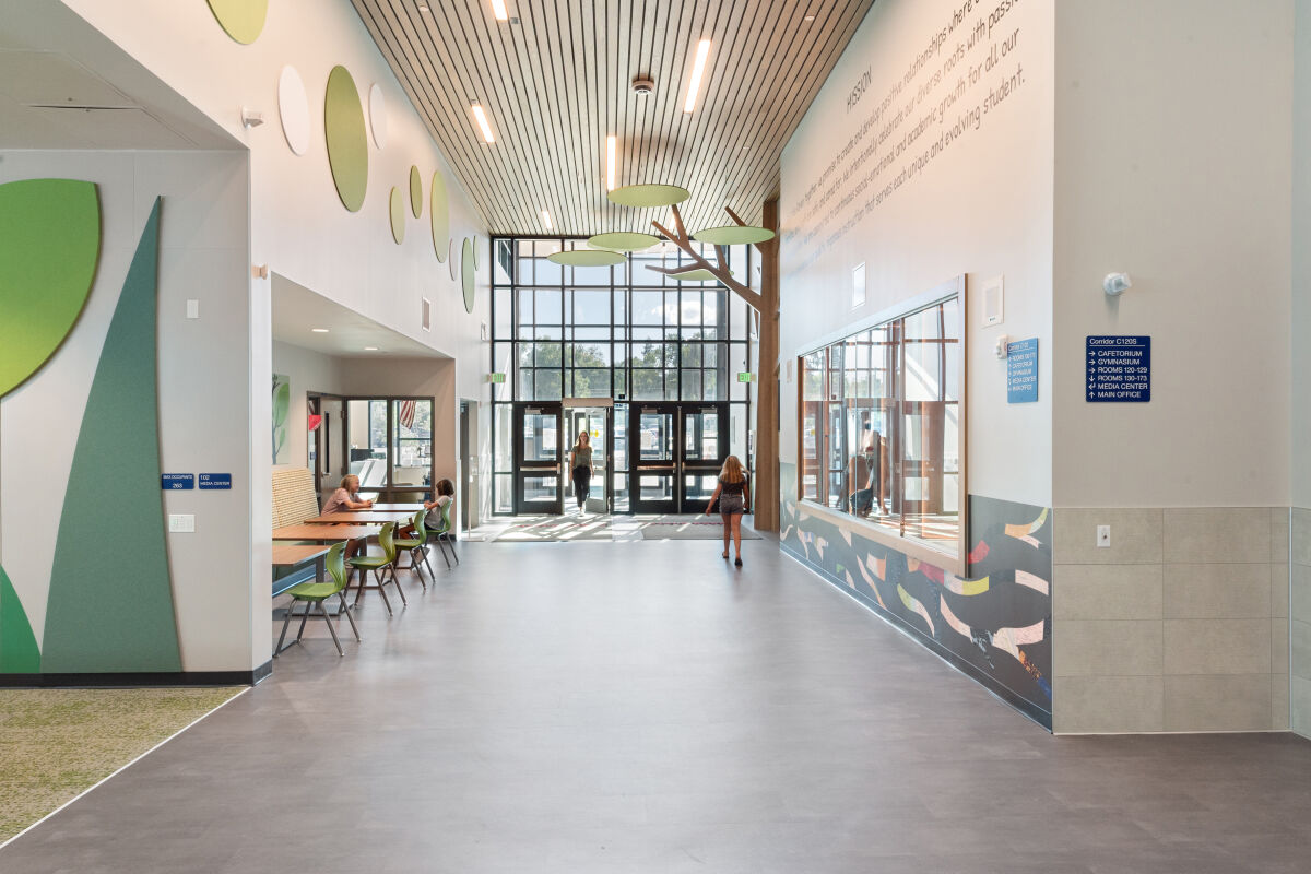 Entry corridor with floor‑to‑ceiling windows at the main doors, large tree‑themed wall installation with circular green canopy panels, leaf‑shaped acoustic wall elements, patterned wall graphics, and adjacent seating alcove with tables and green chairs.