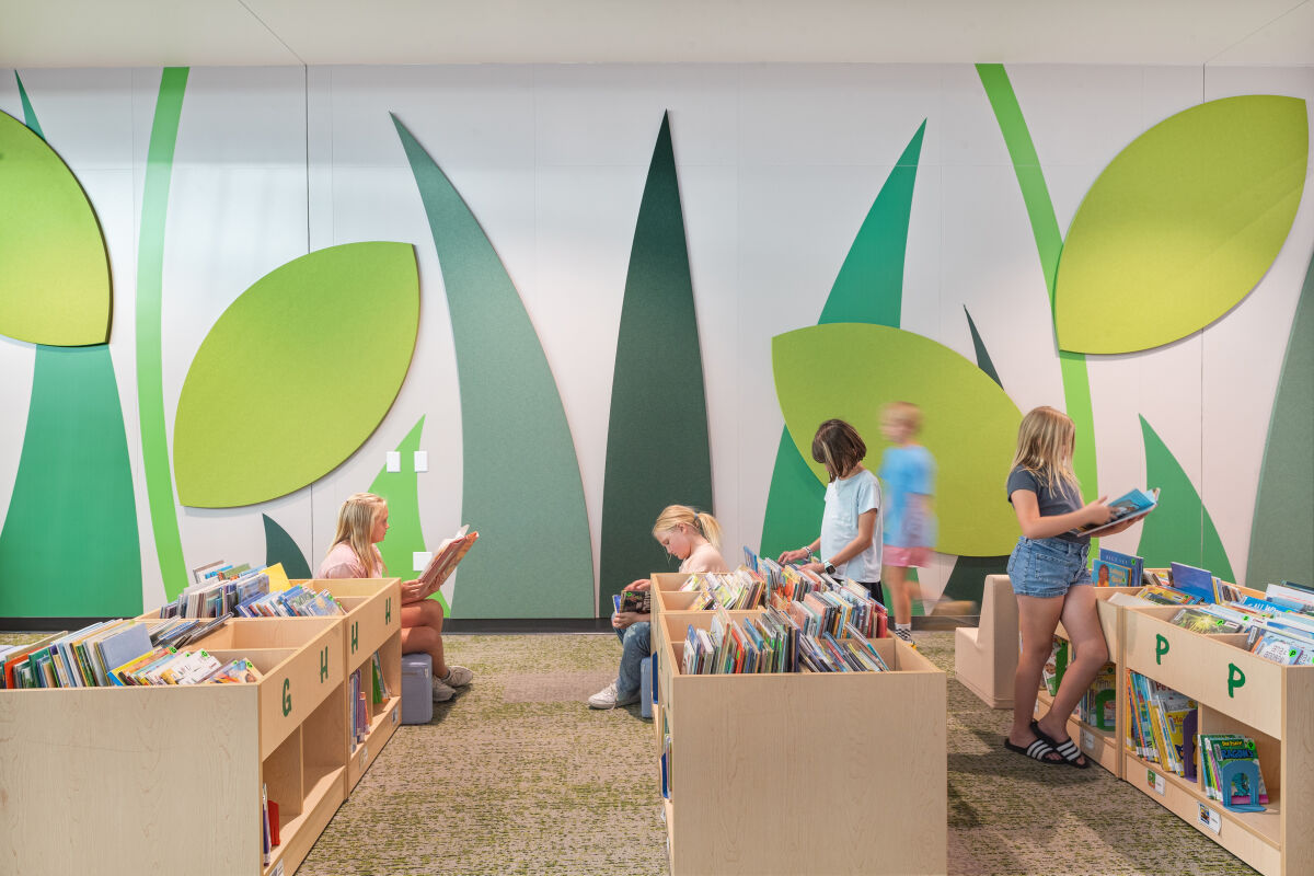 Media center reading area with low book bins organized by alphabet labels, patterned carpet flooring, and a large nature‑themed wall mural featuring oversized green leaf shapes.