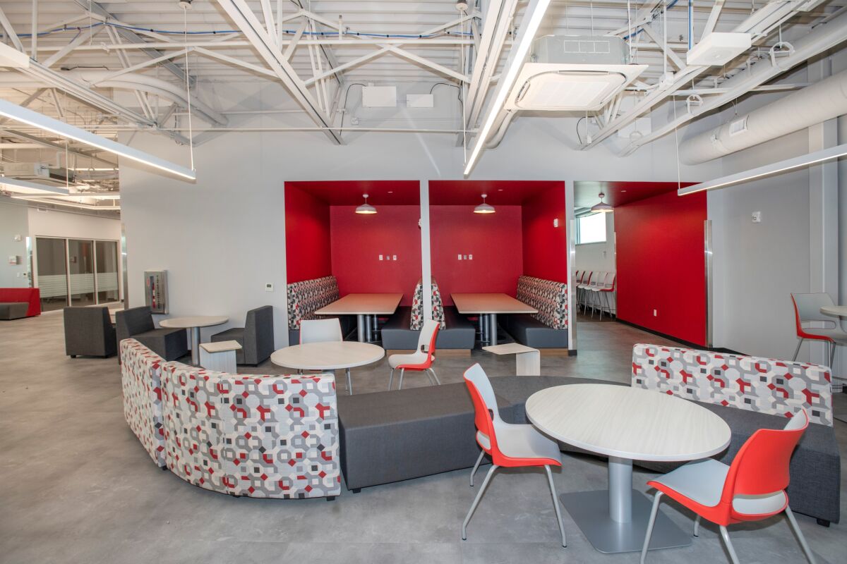 Commons area with patterned lounge seating, round tables and chairs, built‑in red study alcoves with pendant lights, exposed ceiling structure, and polished concrete flooring.