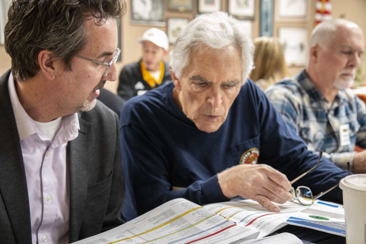 Participants seated at a table reviewing printed documents and charts during a planning meeting, with additional attendees and framed artwork visible in the background.