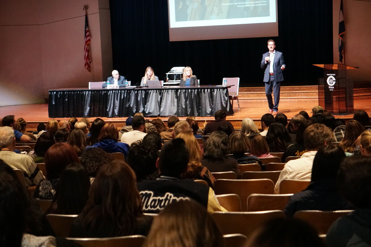 Public meeting in a school auditorium with a presenter speaking at a podium, a panel seated at a table on stage, a projected presentation screen, and attendees seated in rows of auditorium chairs.