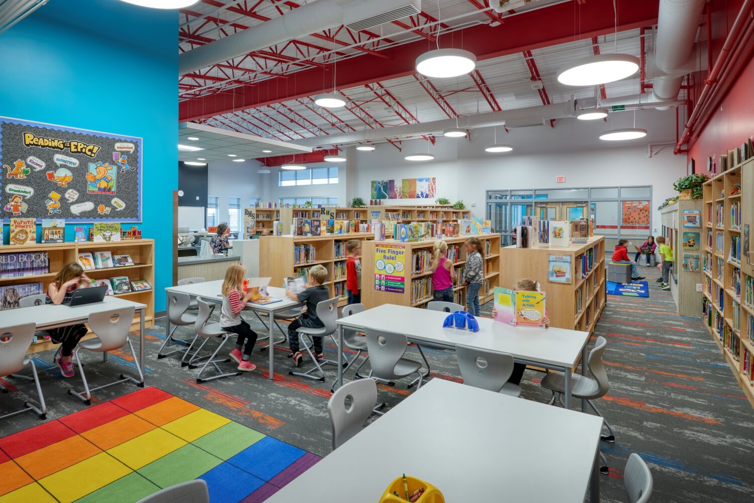 Library with bookshelves, reading tables and chairs, colorful carpet squares, hanging student artwork, and large windows with overhead circular light fixtures.