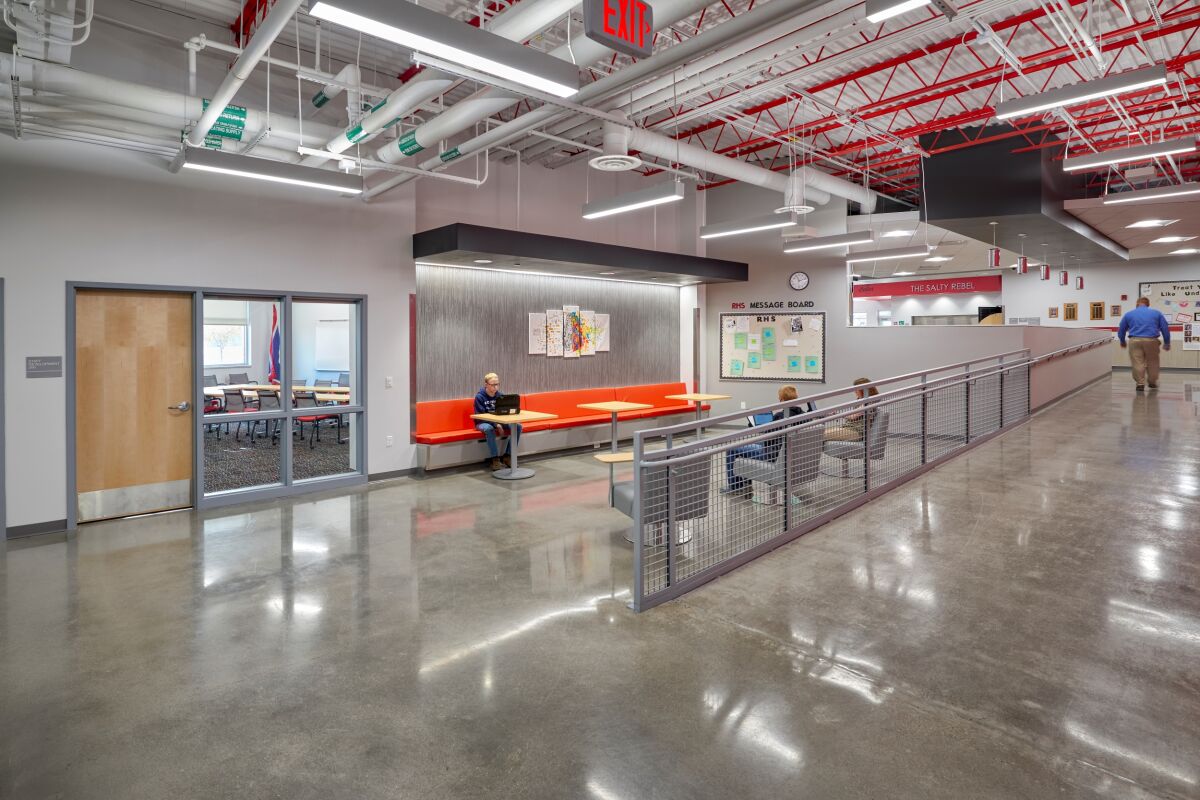 Corridor seating area with an orange bench and small tables in a recessed alcove, polished concrete flooring, exposed ceiling structure with red and white piping, and adjacent classrooms and display boards along the hallway.