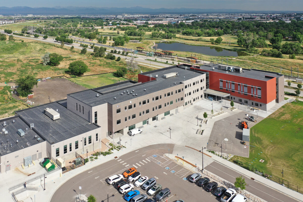 Aerial view of Aurora Science & Technology School with interconnected building wings in gray and red, central courtyard, surrounding parking lot, adjacent open space, and a pond and trail system in the background.