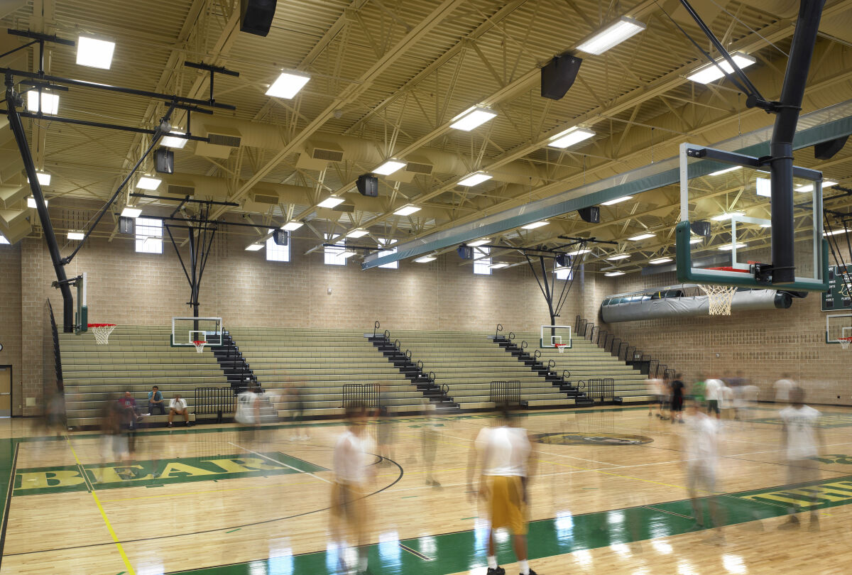 Gymnasium with a hardwood basketball court, retractable bleachers, overhead mechanical systems, multiple basketball hoops, and green and yellow graphics on the floor.