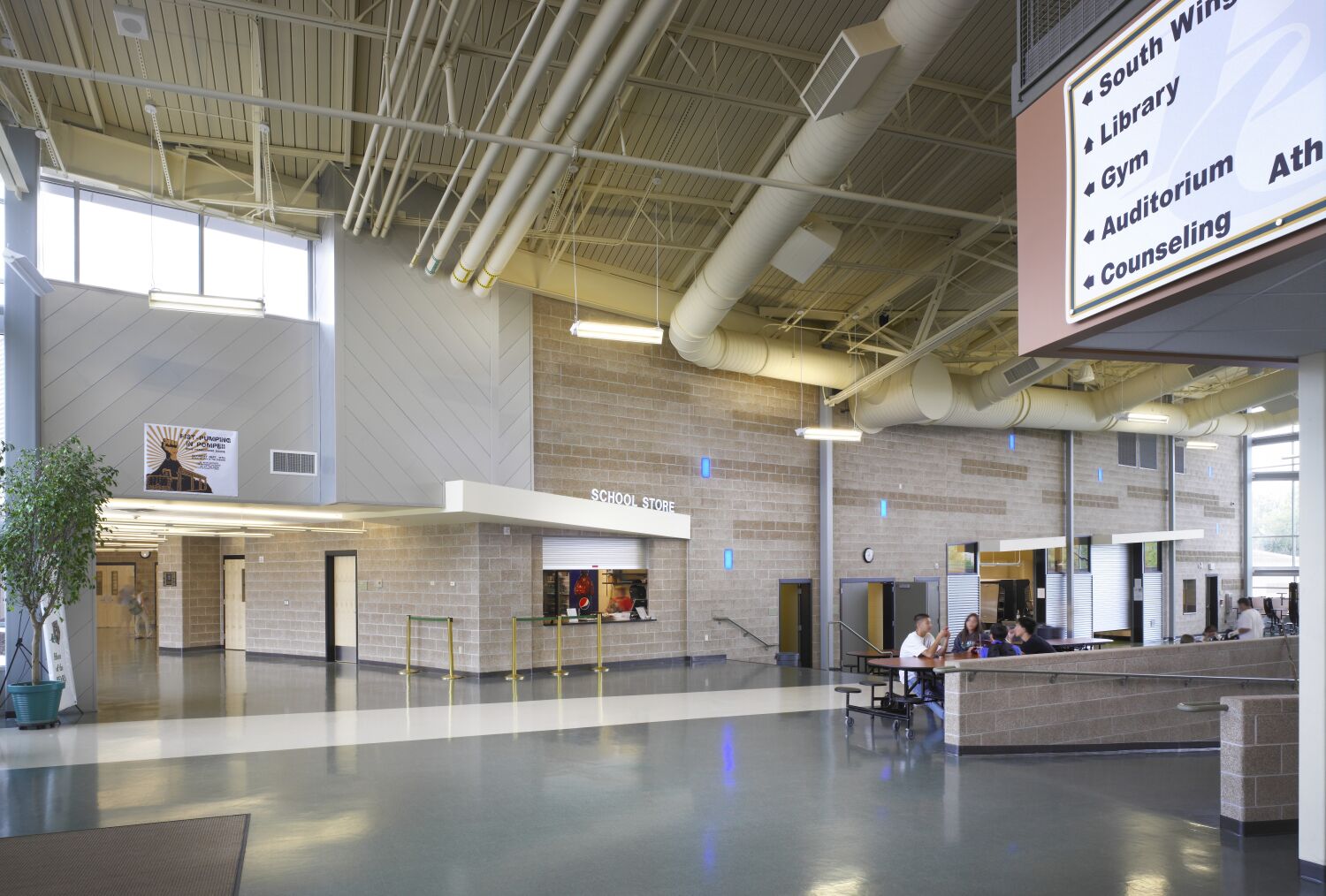Interior commons area with polished flooring, exposed ceiling structure with large ducts, a school store kiosk, signage for building wings, and seating areas along the perimeter.