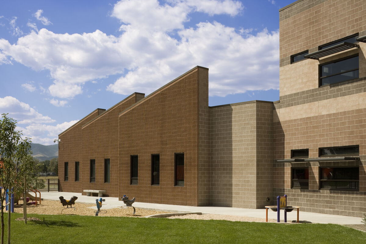 Exterior of Bear Creek K‑8 School with tan and brown brick façades, angled rooflines, small windows, adjacent playground with spring riders, and a grass lawn under a partly cloudy sky.