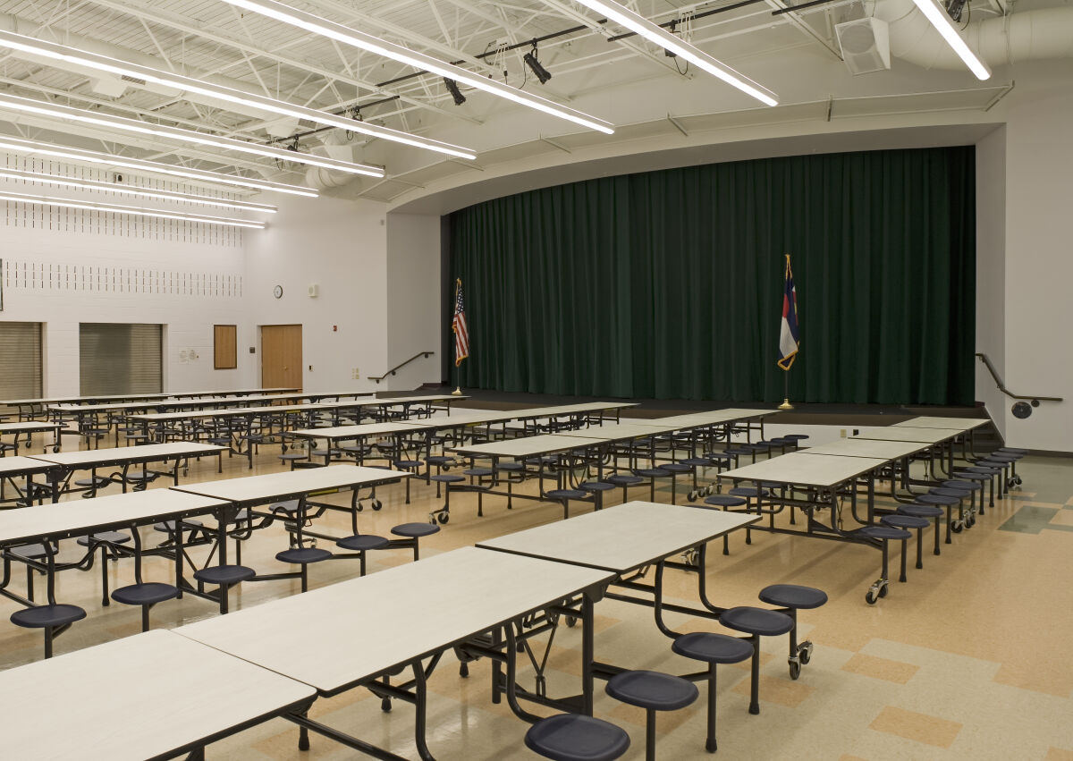 Cafeteria with folding tables and attached stools, polished flooring in neutral tones, and a stage with green curtains beneath an exposed ceiling with linear lighting.