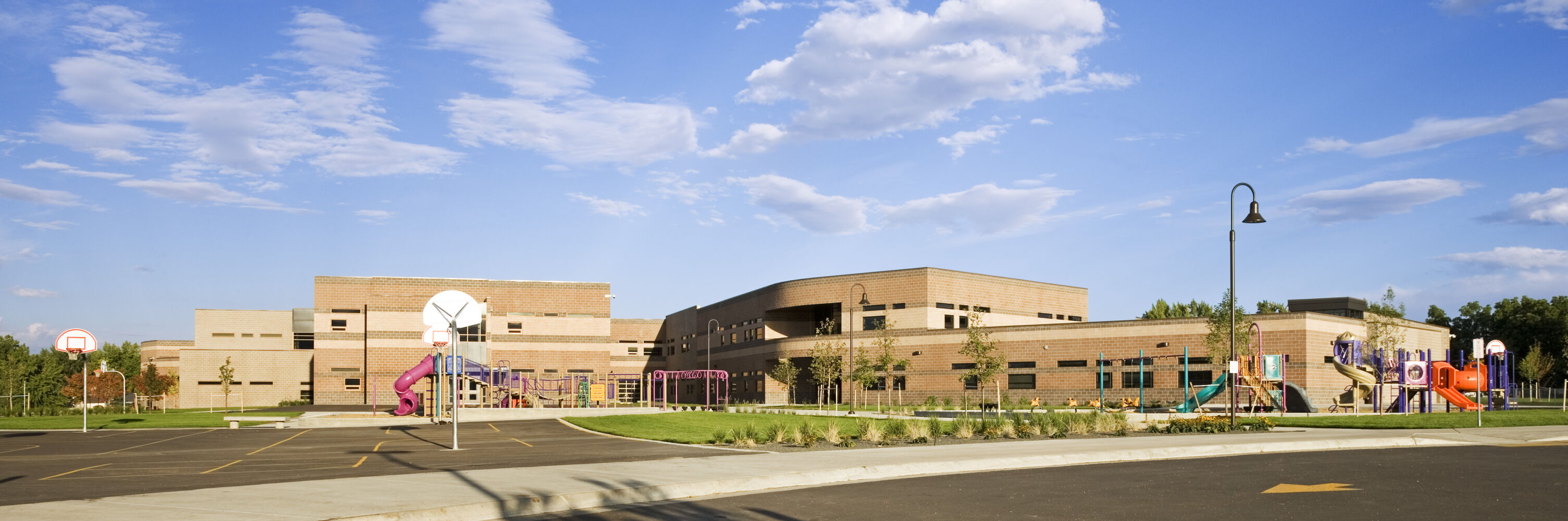 Wide exterior view of Bear Creek K‑8 School with tan and brown brick façades, two playground areas with slides and climbing structures, basketball hoops, and landscaped plantings under a blue sky.