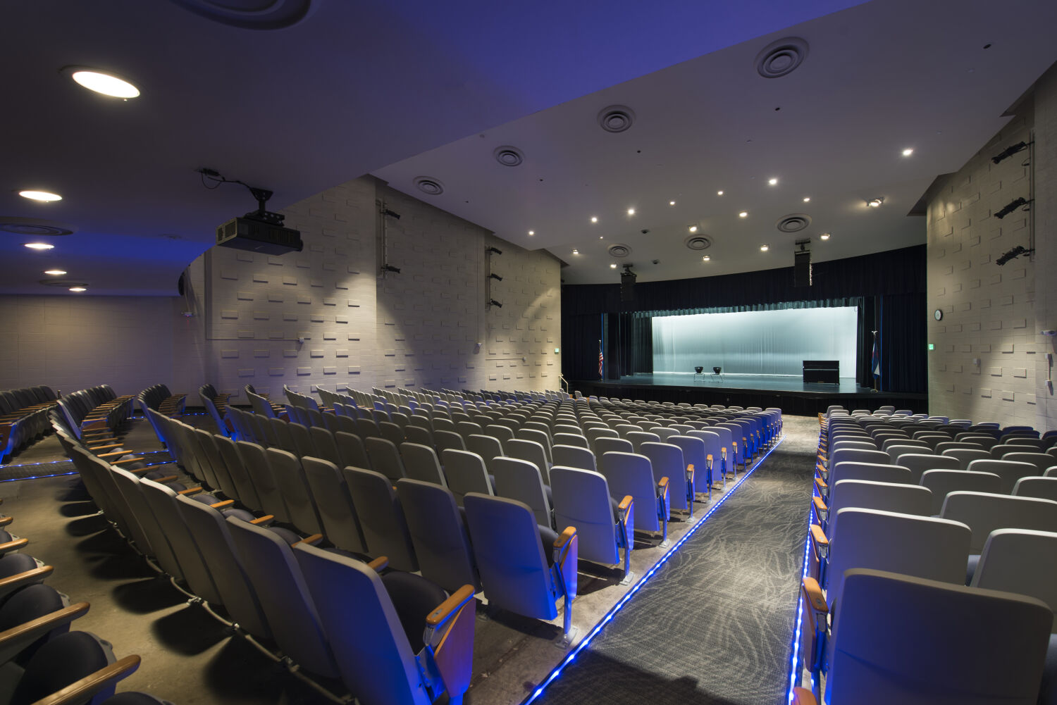 Auditorium with tiered seating, illuminated aisle lighting, textured wall panels, overhead ceiling fixtures, and a proscenium stage with curtains.