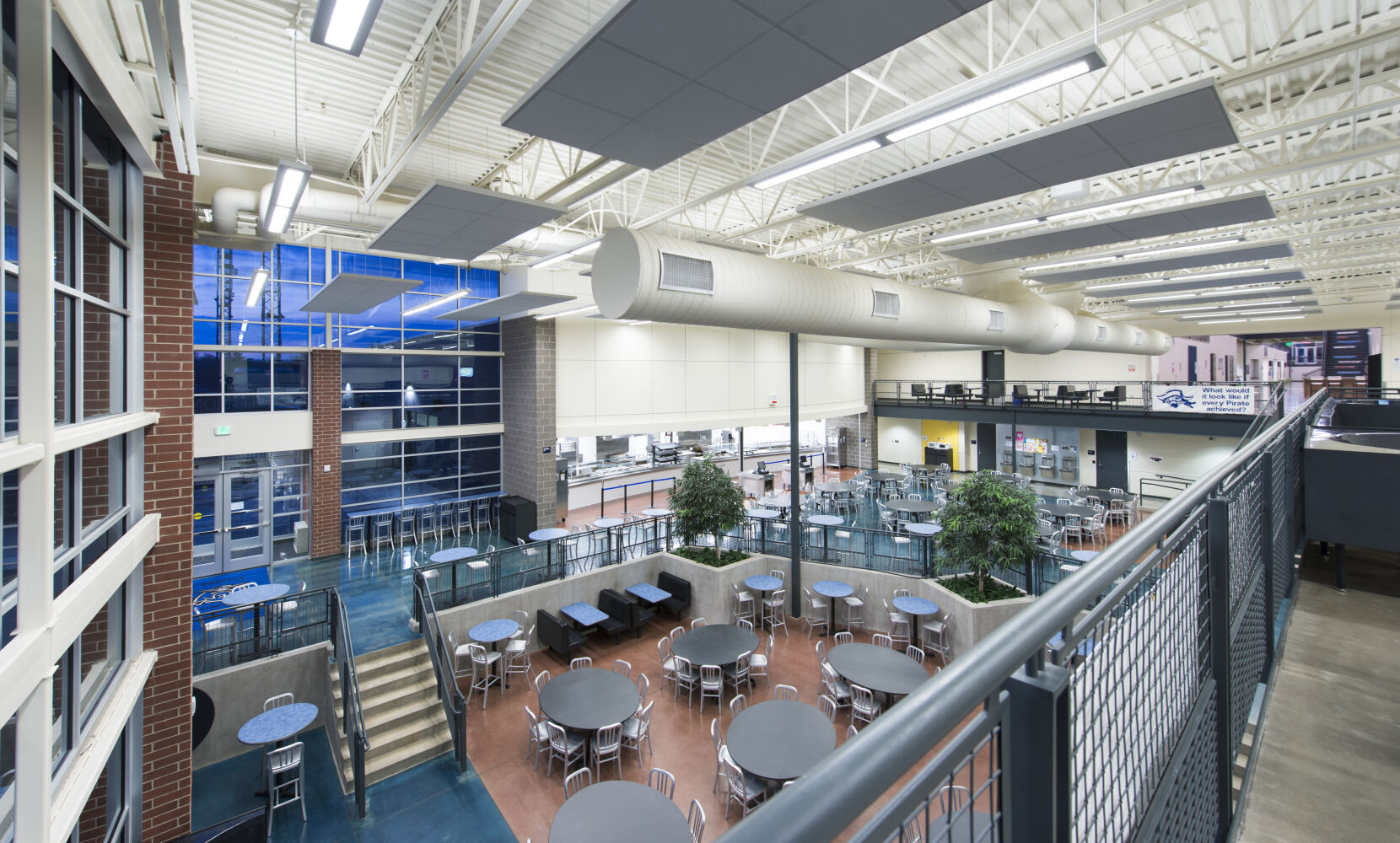 Two‑story cafeteria and commons with round tables, lounge seating, interior trees, metal railings, exposed ceiling with ductwork, and large windows along one wall.
