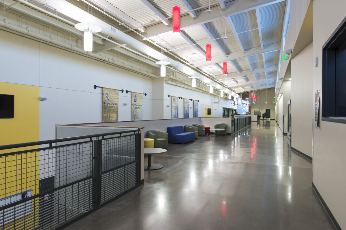 Upper‑level corridor with lounge seating, polished concrete flooring, metal railing overlooking the lower level, colorful pendant lights, and exposed ceiling structure with ductwork and suspended fixtures.