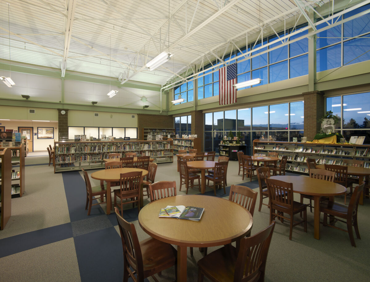 Library with round wooden tables and chairs, bookshelves along the perimeter, large windows with an American flag, carpet tile flooring, and an exposed white truss ceiling with suspended lighting.