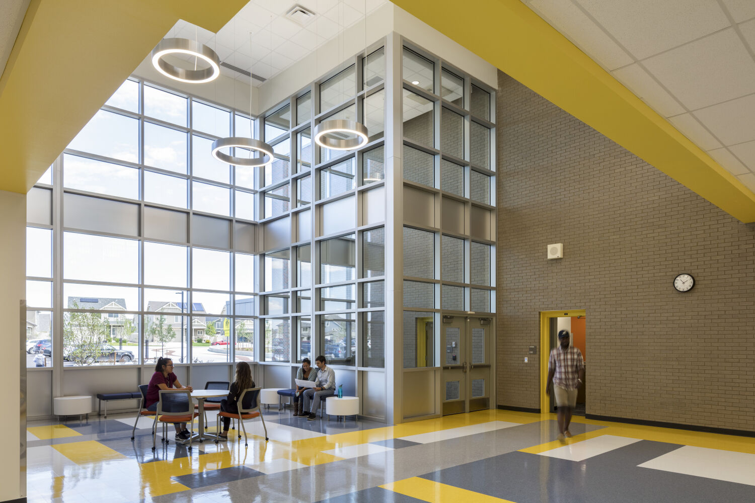 Two‑story commons area with large floor‑to‑ceiling windows, circular pendant lights, yellow and gray accent walls, patterned tile flooring, and small seating tables near the glass wall.