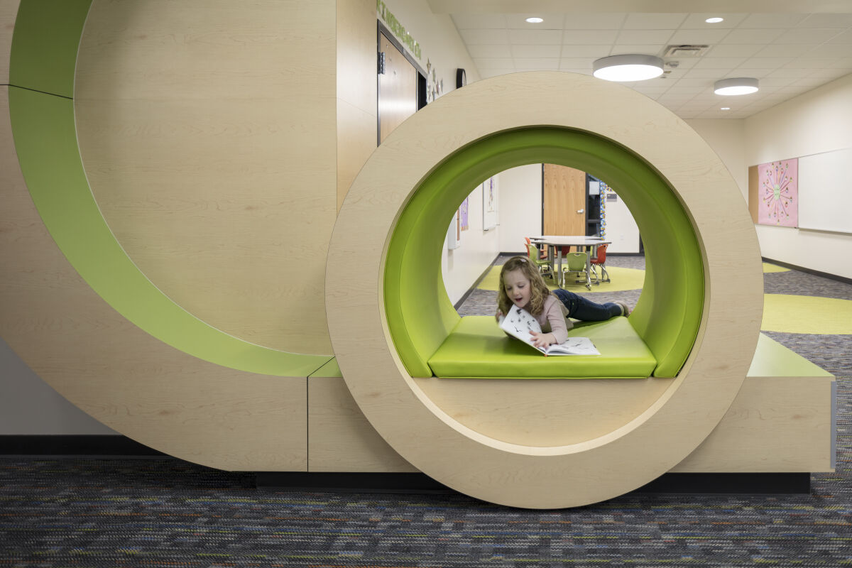 Circular built‑in reading nook with green padded seating set into a wood‑paneled wall, located along a classroom corridor with patterned carpet and adjacent learning spaces.