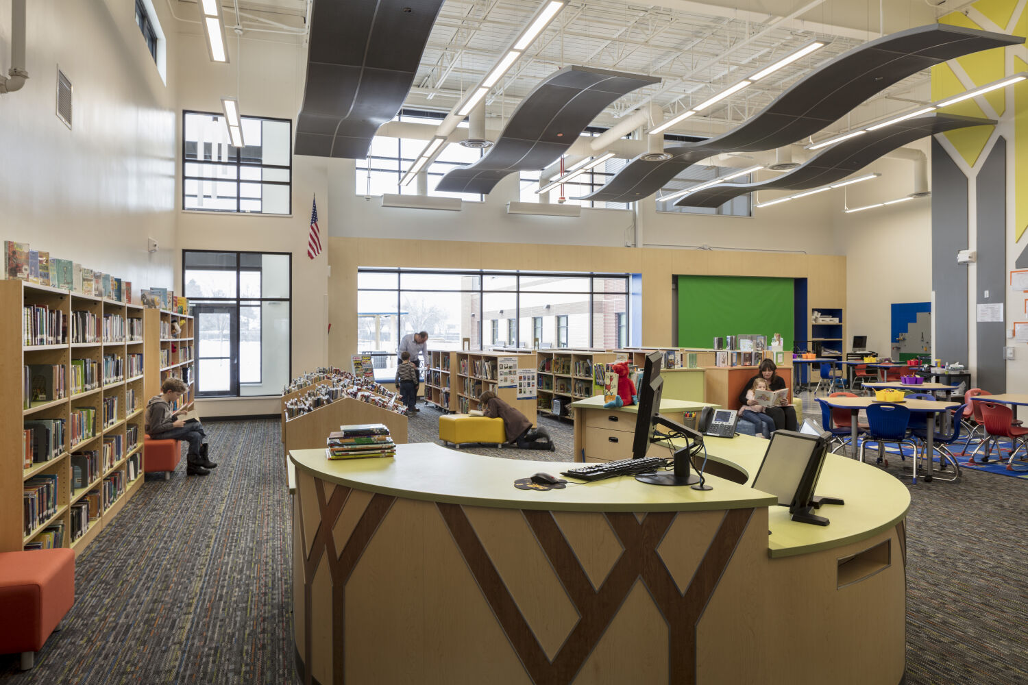 Library and media center with curved circulation desk, perimeter bookshelves, large windows, patterned carpet flooring, movable seating, a green screen wall, and overhead curved acoustic panels.
