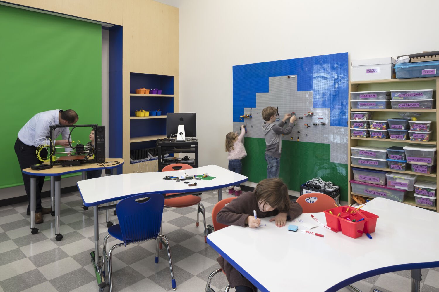 Makerspace with a green‑screen wall, LEGO building panel, storage shelves with labeled bins, white activity tables with blue and red chairs, and a workstation with a 3D printer.