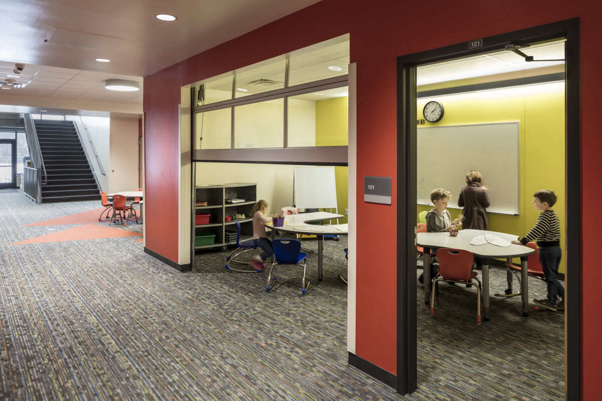Small‑group learning rooms with red accent walls, whiteboards, student tables and chairs, open shelving with bins, and a corridor with patterned carpet and a central staircase.