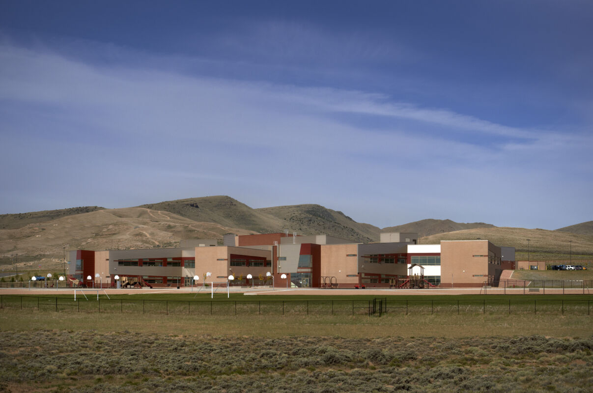 Distant exterior view of Rawlins Elementary School with multiple connected brick and metal‑panel building volumes, outdoor basketball courts, and surrounding open landscape with rolling hills.