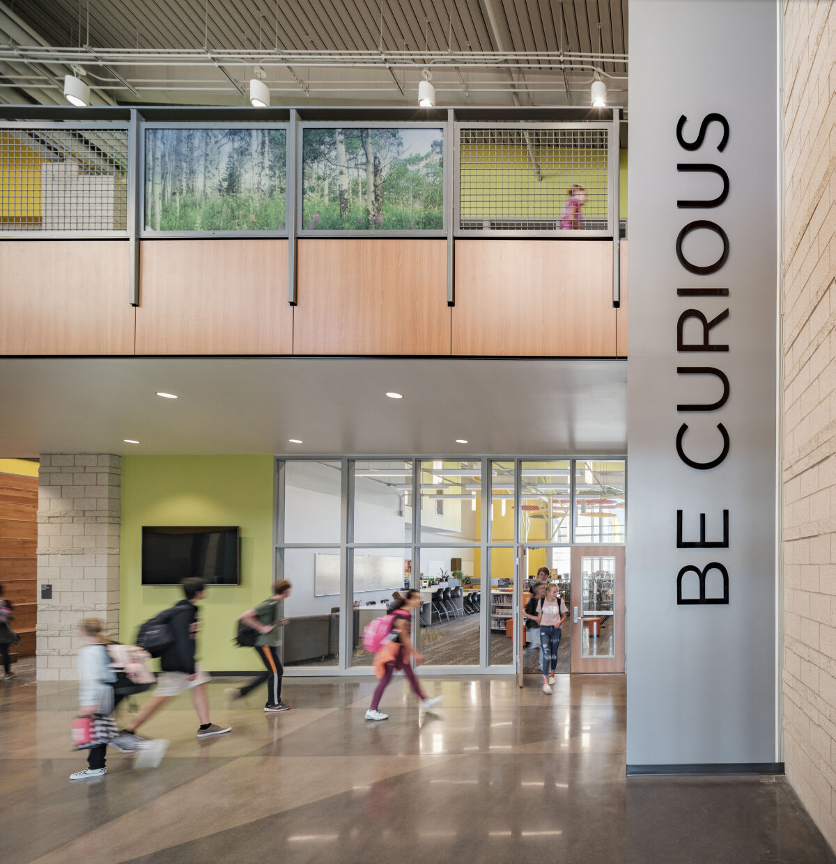 Two‑story corridor with a tall ‘BE CURIOUS’ wall graphic, mesh‑panel balcony railing with nature imagery, large windows, polished concrete flooring, and adjacent classrooms with glass walls.