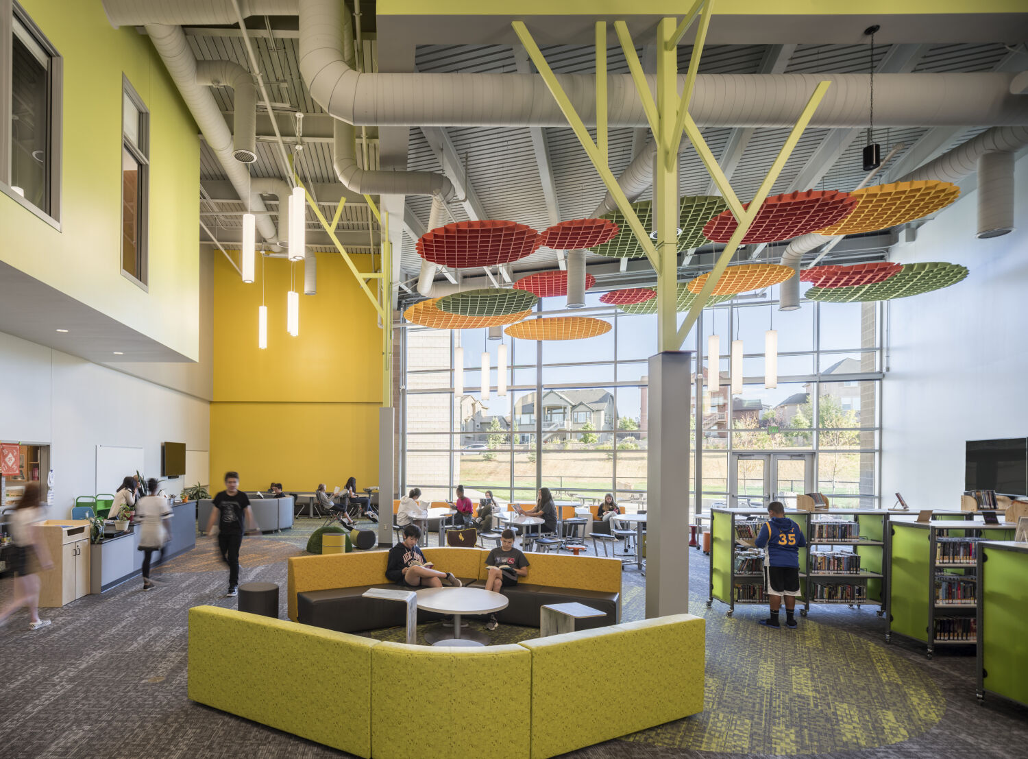 Learning commons with colorful overhead acoustic panels, tree‑like structural columns, round lounge seating, bookshelves, café‑style tables near large windows, and exposed ceiling ductwork.