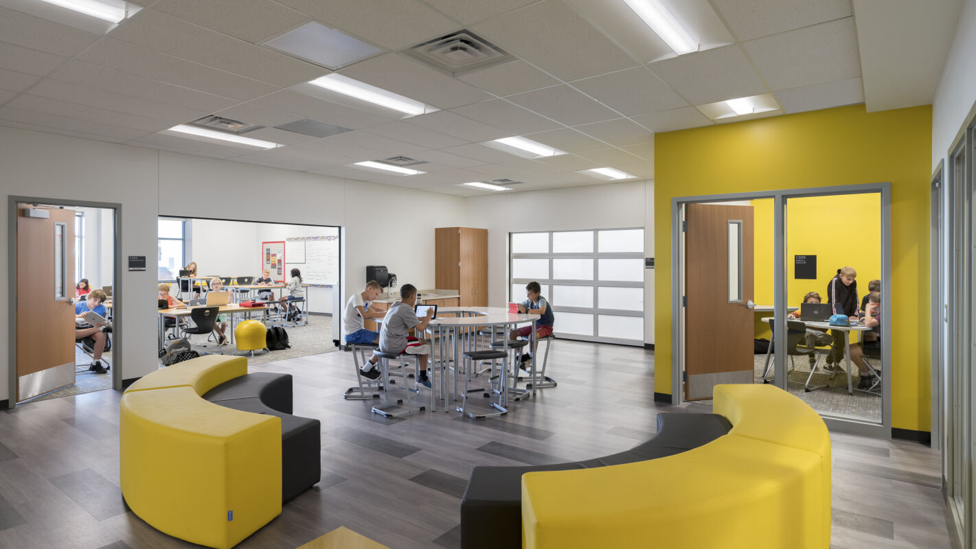 Small‑group learning suite with yellow accent walls, curved lounge seating, high‑top tables and stools, adjacent classrooms with glass partitions, and patterned flooring.