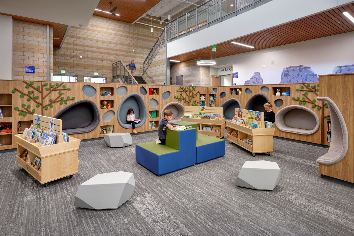 Media center reading area with built‑in tree‑themed shelving, pod‑style reading nooks, movable book carts, soft seating blocks, and patterned carpet within a two‑story corridor.