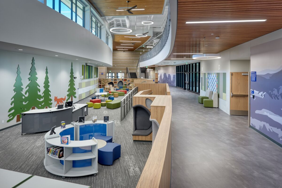 Media center with forest‑themed wall graphics, curved wood ceiling panels, circular pendant lights, built‑in reading nooks, movable soft seating, bookshelves, and large upper windows along a two‑story corridor.