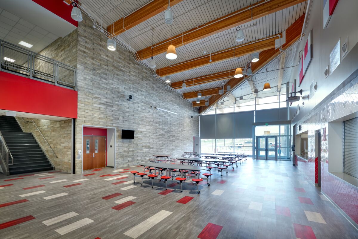 The Deer Trail PK–12 School cafeteria commons featuring a sloped wood ceiling, large windows, stone accent wall, red and gray flooring patterns, cafeteria tables, and an adjacent stairway.