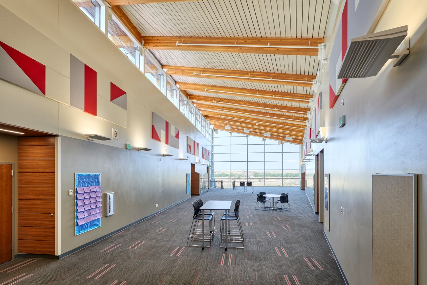 Interior hallway of Deer Trail PK–12 School featuring a sloped wood ceiling, natural light from a full-height glass wall, lockers, and seating along the corridor.