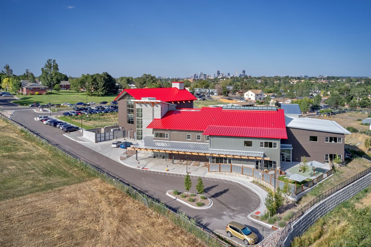 Exterior view of Great Work Montessori PK–5 School showing the glass entry tower, red rooflines, wood canopy structure, vertical siding, parking area, and surrounding landscaping under a partly cloudy sky.