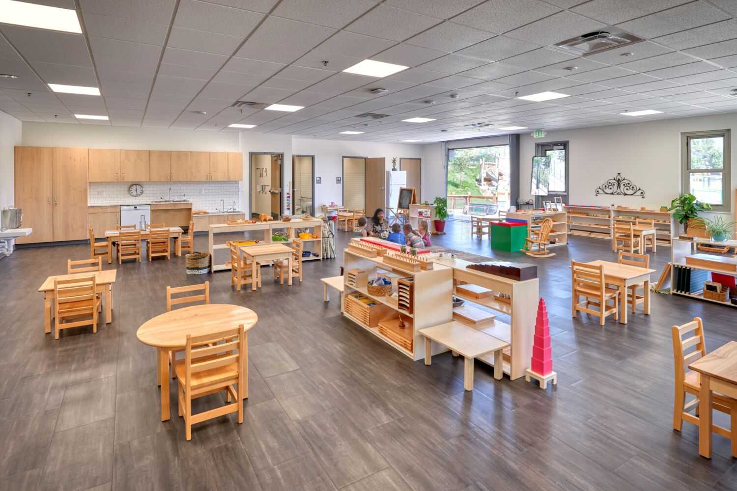 Great Work Montessori PK–5 School classroom featuring low wooden tables and chairs, open shelving with Montessori learning materials, built‑in cabinetry and sinks, large windows for natural light, and children working at activity stations.