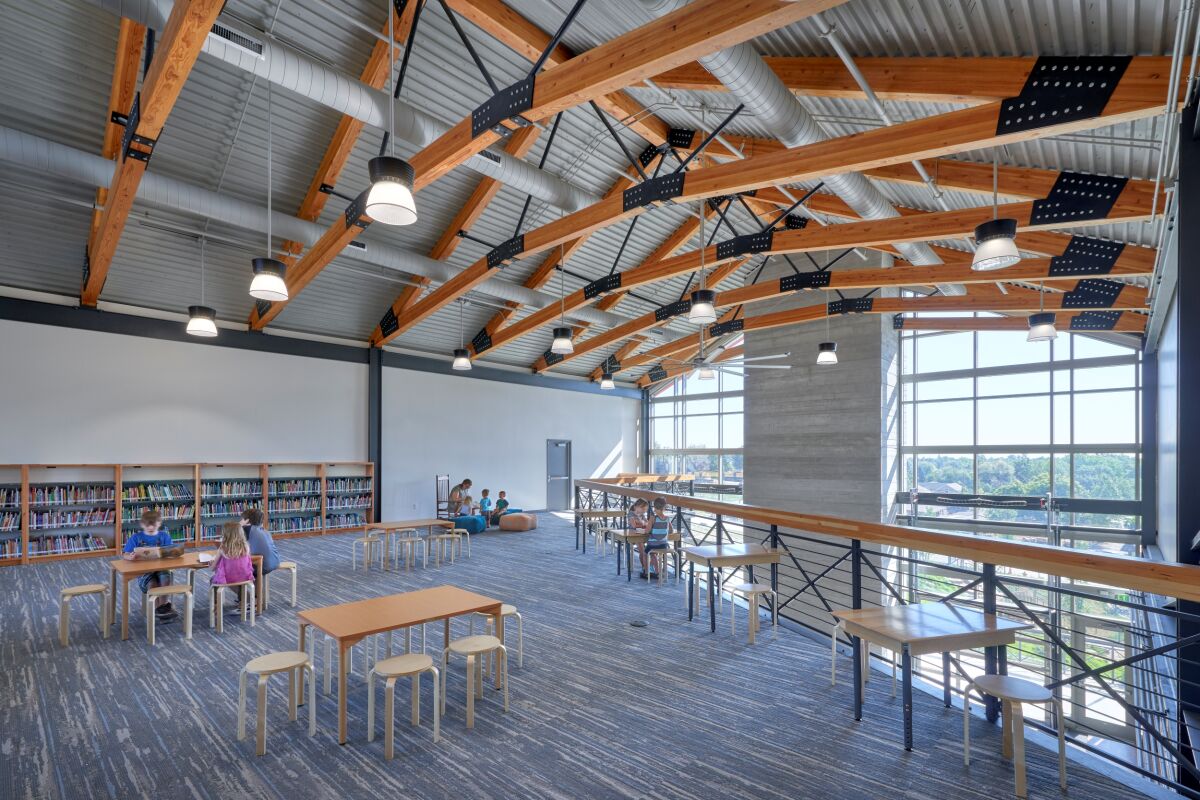 Great Work Montessori PK–5 School library loft featuring exposed wood and steel trusses, tall glass windows overlooking the landscape, bookshelves along the wall, and tables with chairs arranged on an area rug.