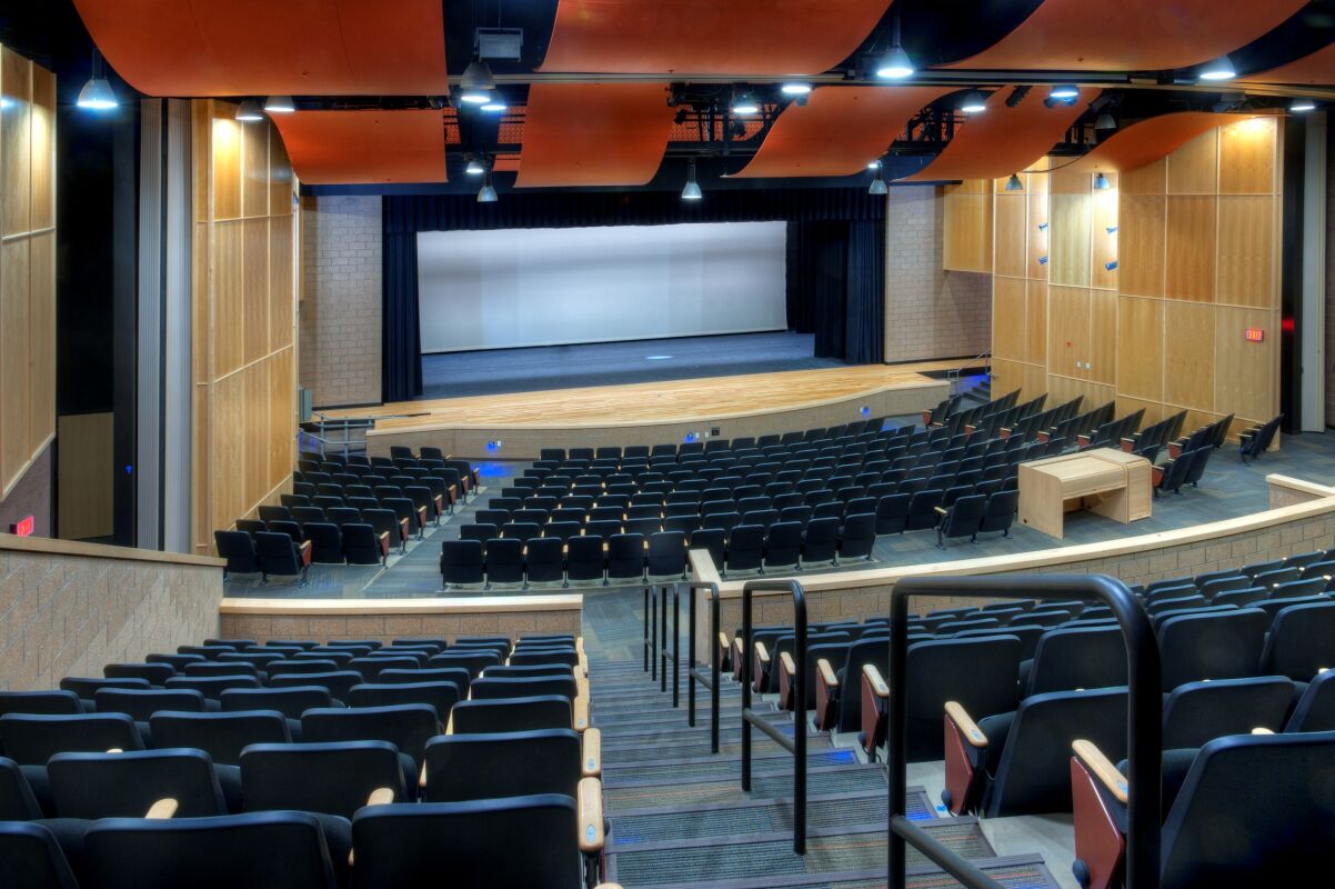 Auditorium showing tiered seating, a large performance stage with a curtain, wood wall paneling, vertical acoustic panels, and curved ceiling baffles with integrated lighting.