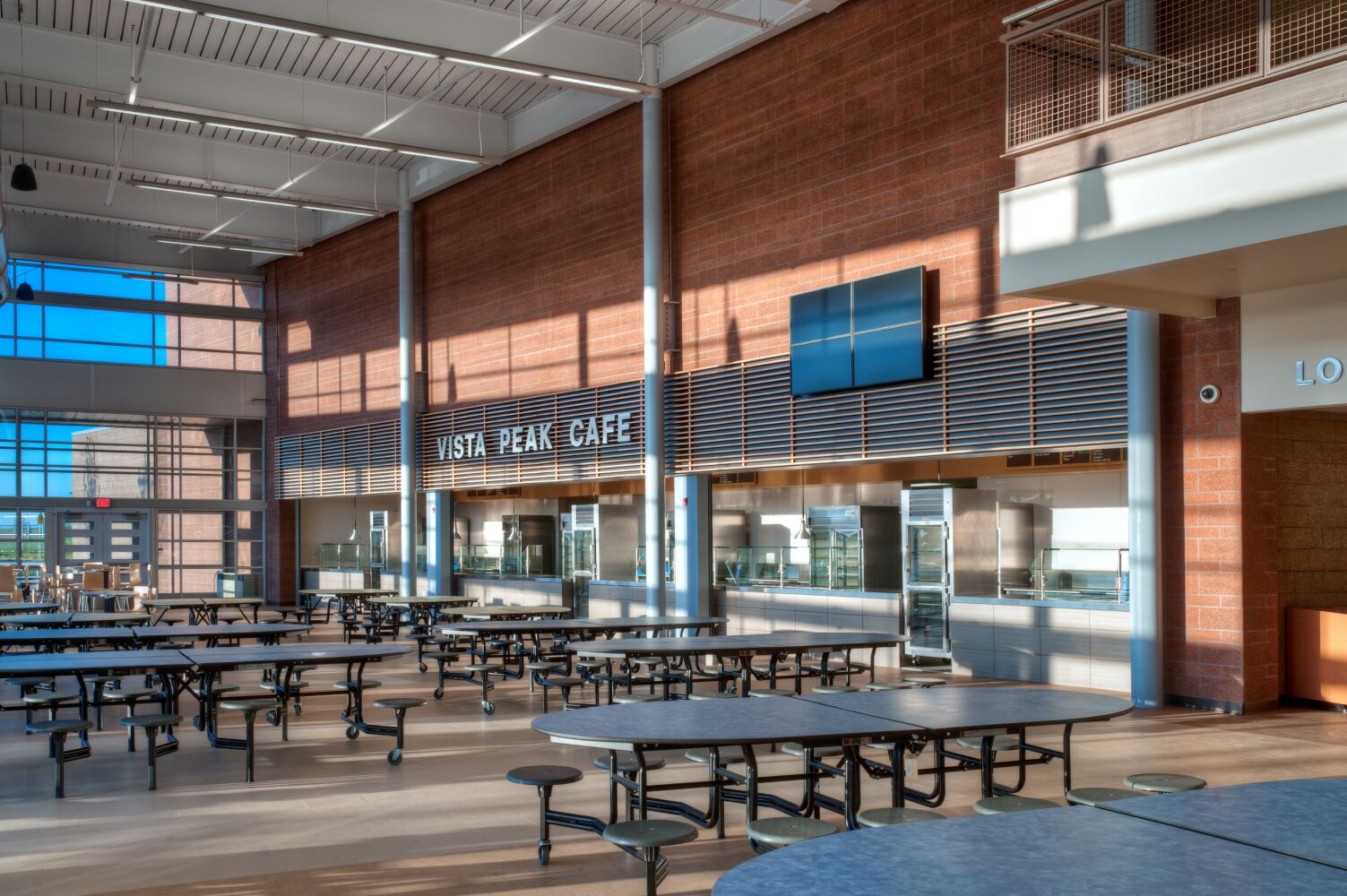 Cafeteria featuring round cafeteria tables, tall glass windows, a brick accent wall with the Vista Peak Café serving stations, overhead pendant lighting, and an open, double-height space filled with natural light.