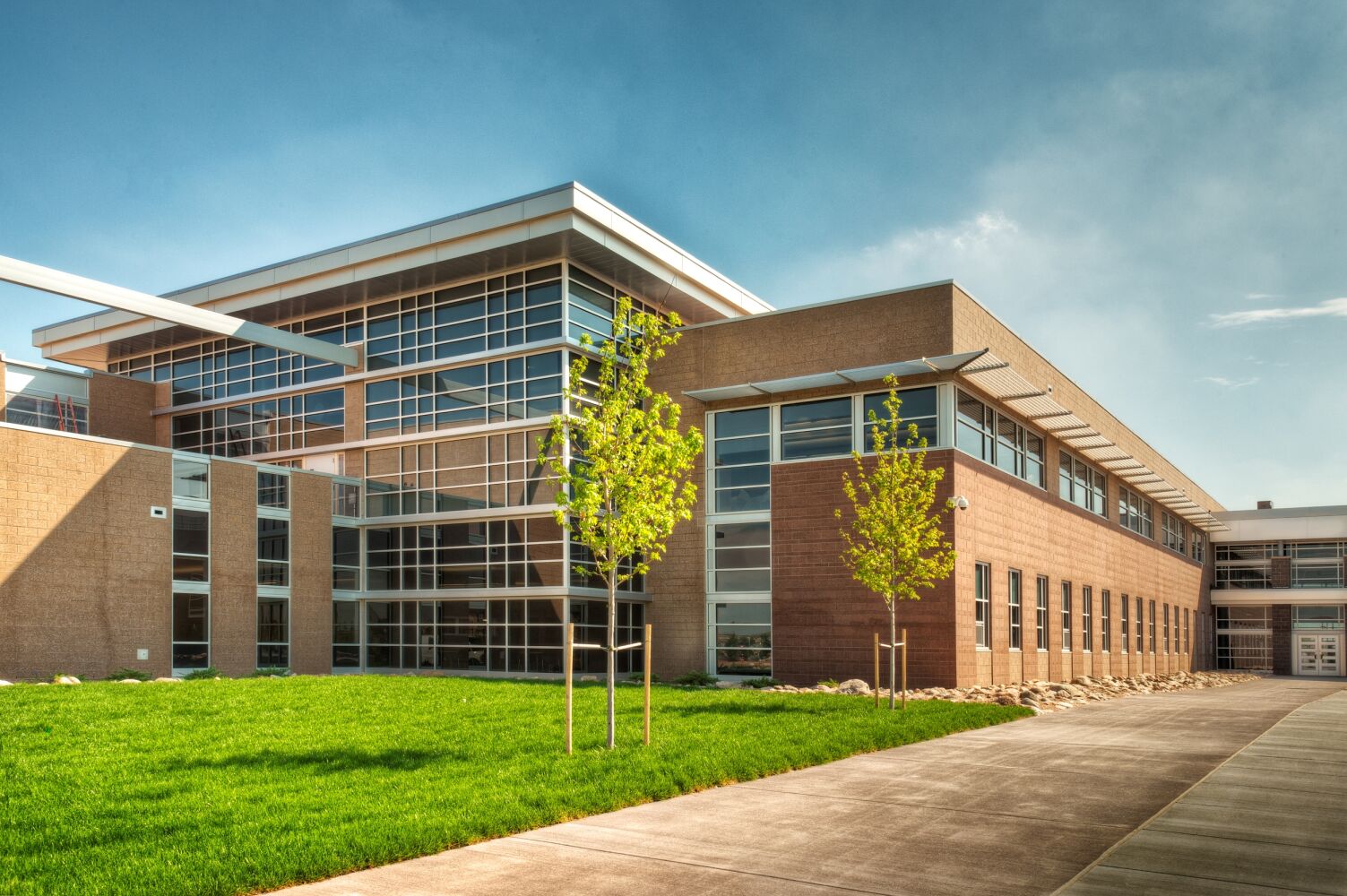Classroom wing featuring large glass curtainwall windows, brick façades, a landscaped lawn with young trees, and a concrete walkway under a clear sky.