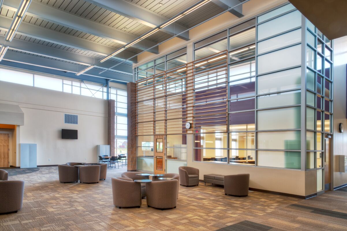 Learning commons featuring lounge seating, a patterned carpeted floor, large glass partition walls with horizontal wood slats, high ceilings with exposed structure, and tall windows bringing in natural light.