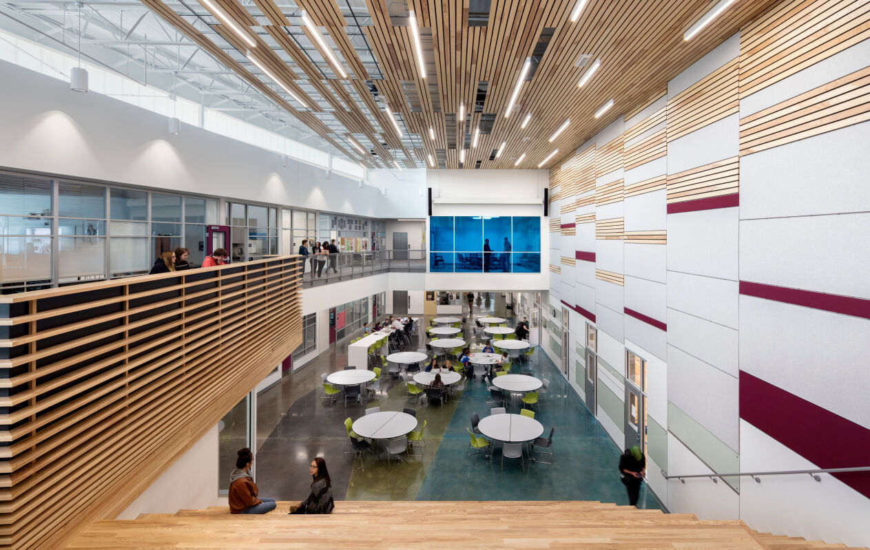 Pathways Innovation Center learning commons featuring wide tiered wood stairs, round tables with chairs on polished concrete flooring, a glass-walled upper walkway, and a ceiling of suspended wood slats with linear LED lighting.