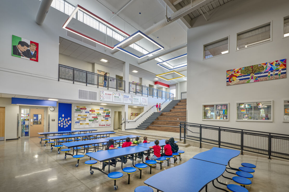 View of the cafeteria commons with blue tables, wide wood learning stairs, a mezzanine walkway, geometric ceiling lights, and colorful wall panels.