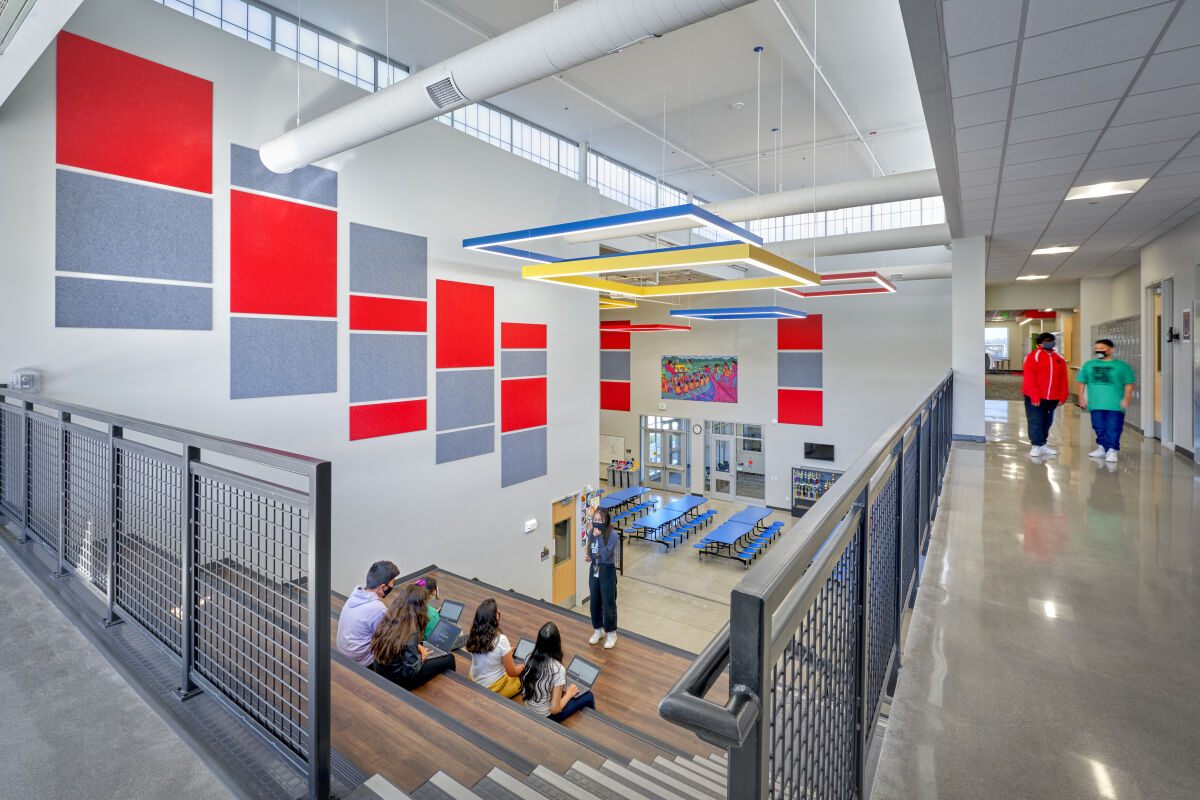 Learning stairs and cafeteria commons with colorful wall panels, geometric ceiling lights, metal railings, polished concrete floors, and high clerestory windows.