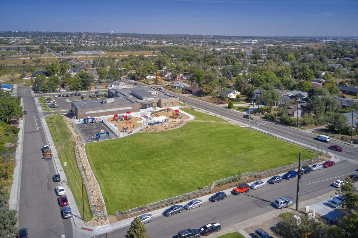 Aerial view of Ricardo Flores Magón Academy showing the main school building, large green open field, multiple playground areas with play structures, and a wide view of the surrounding neighborhood and landscape.