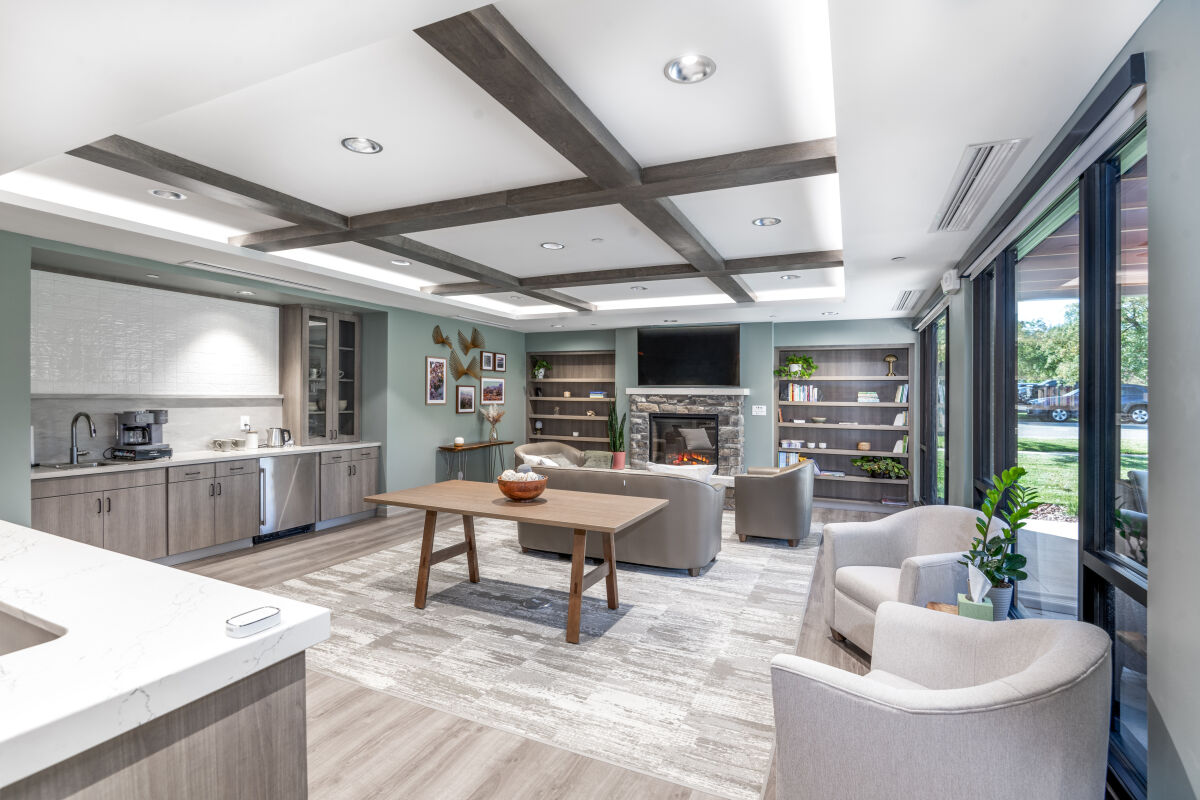 Warm living room lobby with a stone fireplace, built‑in shelving, gray lounge seating, wood ceiling beams, large windows, and a kitchenette with light wood cabinetry.