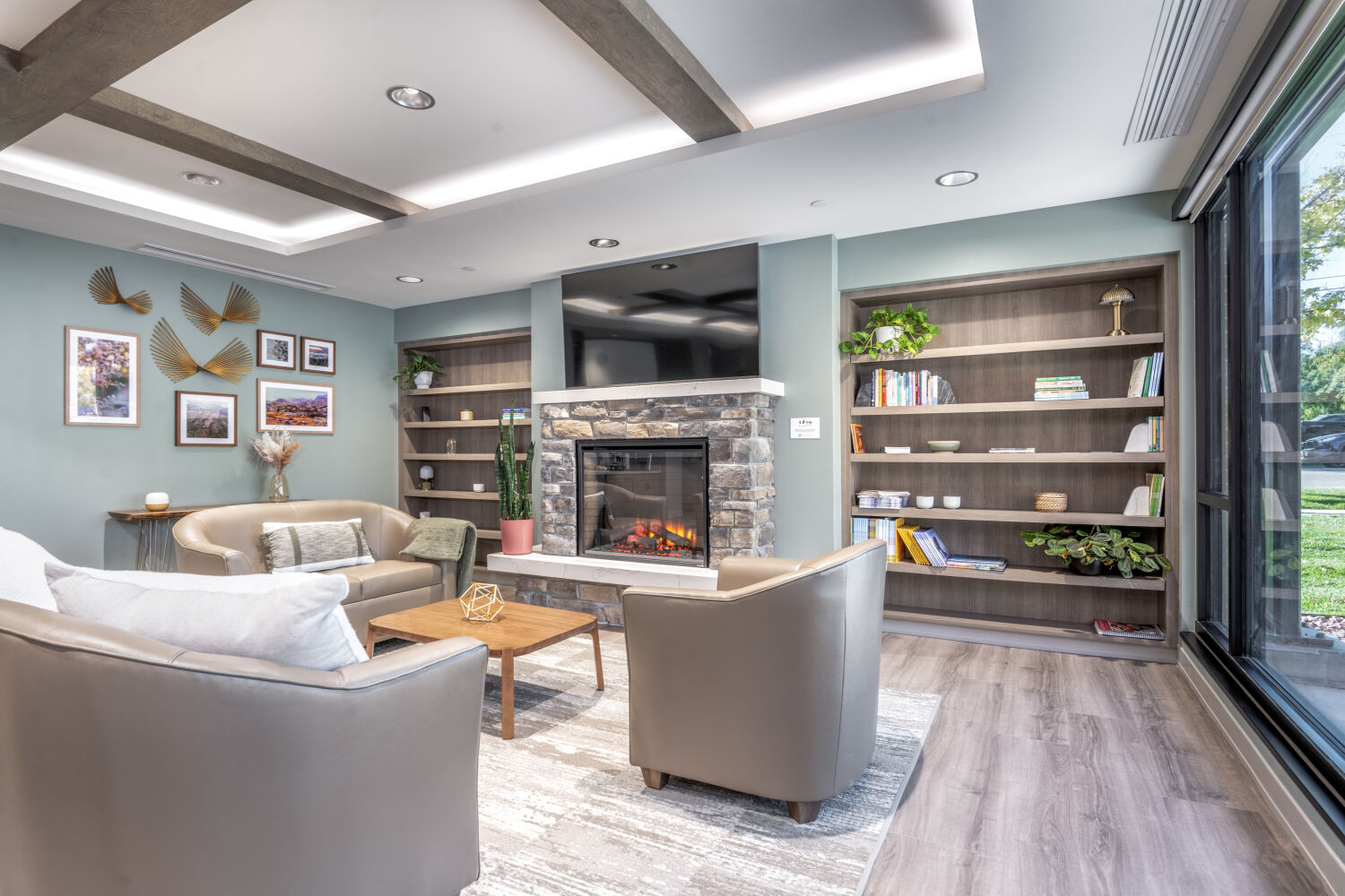 Waiting room with a stone fireplace, built-in wood shelving, framed artwork, neutral upholstered seating, and large windows overlooking the landscape.