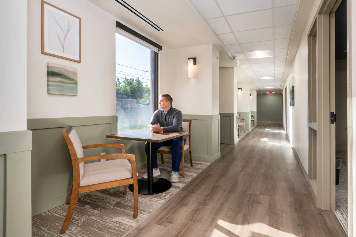 Respite area along a corridor with upholstered armchairs, a small wood table, pale green wainscoting, framed artwork, and a large window bringing in natural light.