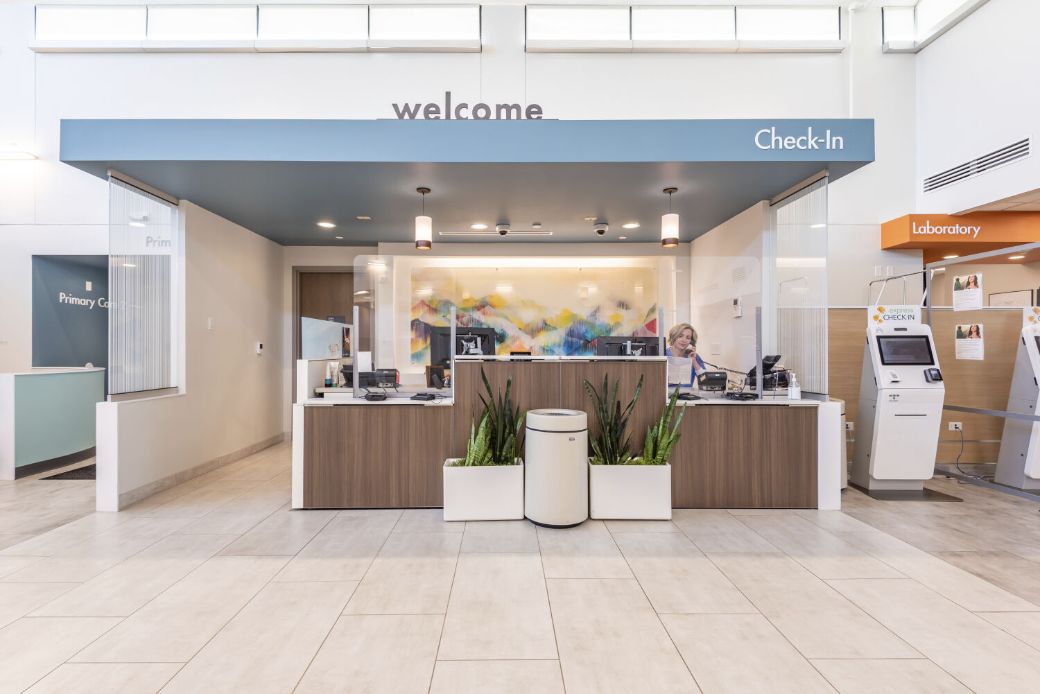 Check-in desk with a blue welcome canopy, wood-front counters, potted plants, tiled flooring, and a colorful abstract mural behind the workstation area.