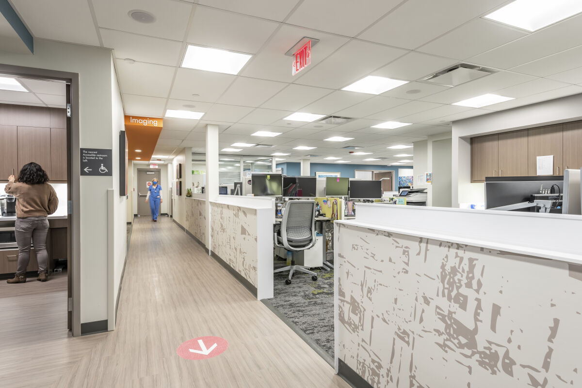 Clinic workspace with open workstations, wood cabinetry, a side kitchenette, overhead lighting, wood-look flooring, and a corridor leading to the imaging area.
