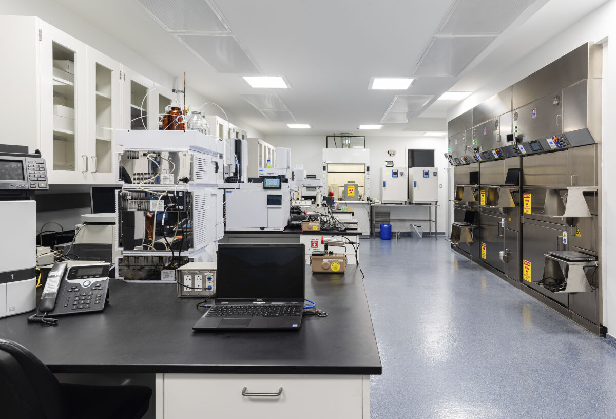 Radiopharmacy lab with stainless steel hot cells, analytical instruments along black countertops, upper storage cabinets, fume hood, epoxy flooring, and overhead fluorescent lighting.
